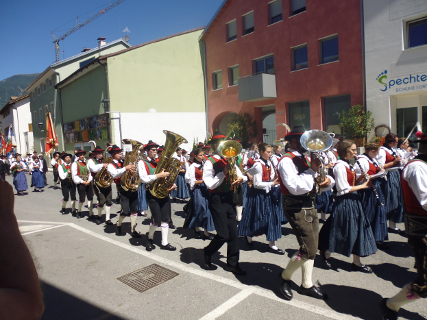 Eine Gruppe von Menschen in traditioneller bayrischer Tracht, die auf der Straße musizieren, während sie durch eine Straße mit Gebäuden gehen, einige halten Fahnen, mit einem Hügel und einem klaren blauen Himmel im Hintergrund.