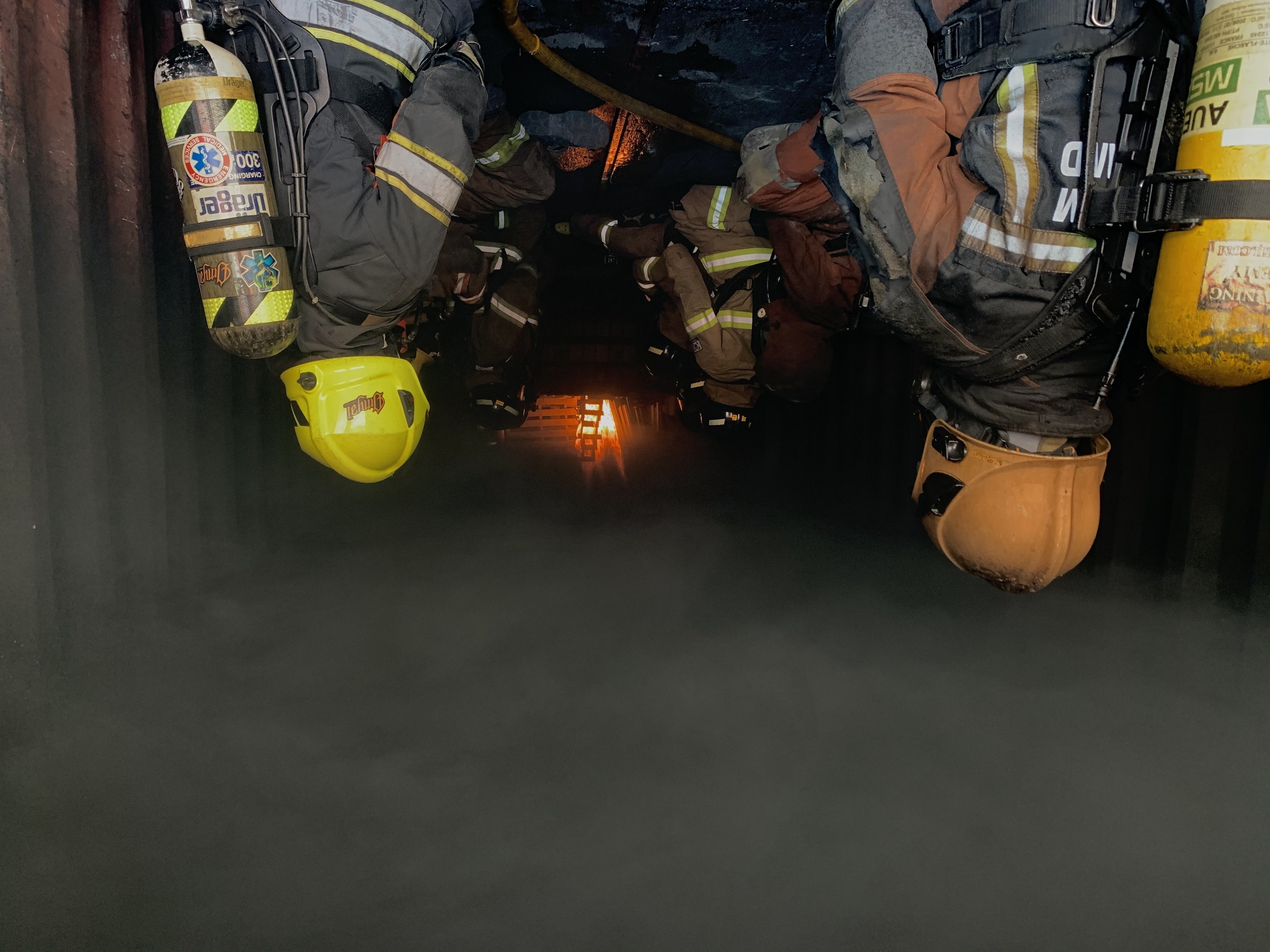 Feuerwehrleute in Schutzausrüstung und Atemschutz arbeiten in einem raucherfüllten Tunnel mit einer Lichtquelle im Hintergrund.