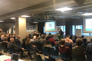 Eine große Gruppe von Menschen sitzt auf Stühlen und schaut auf eine Projektionswand, daneben steht ein Redner mit einem Mikrofon und einem Gerät auf einem Ständer in einem Raum mit Säulen, Fenstern, Deckenleuchten und einer Tafel mit Text.