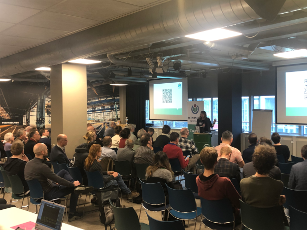 Eine große Gruppe von Menschen sitzt auf Stühlen und schaut auf eine Projektionswand, daneben steht ein Redner mit einem Mikrofon und einem Gerät auf einem Ständer in einem Raum mit Säulen, Fenstern, Deckenleuchten und einer Tafel mit Text.