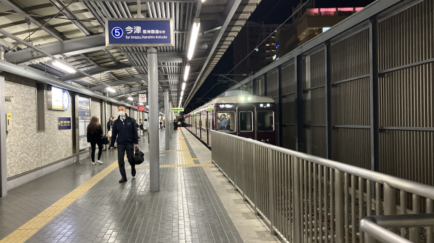 Ein Zug fährt auf ein Bahnsteiggelände mit Menschen in der Nähe, einem Geländer auf der rechten Seite, Gebäuden im Hintergrund, Bahnsteigbeleuchtung und einer Anzeigetafel.