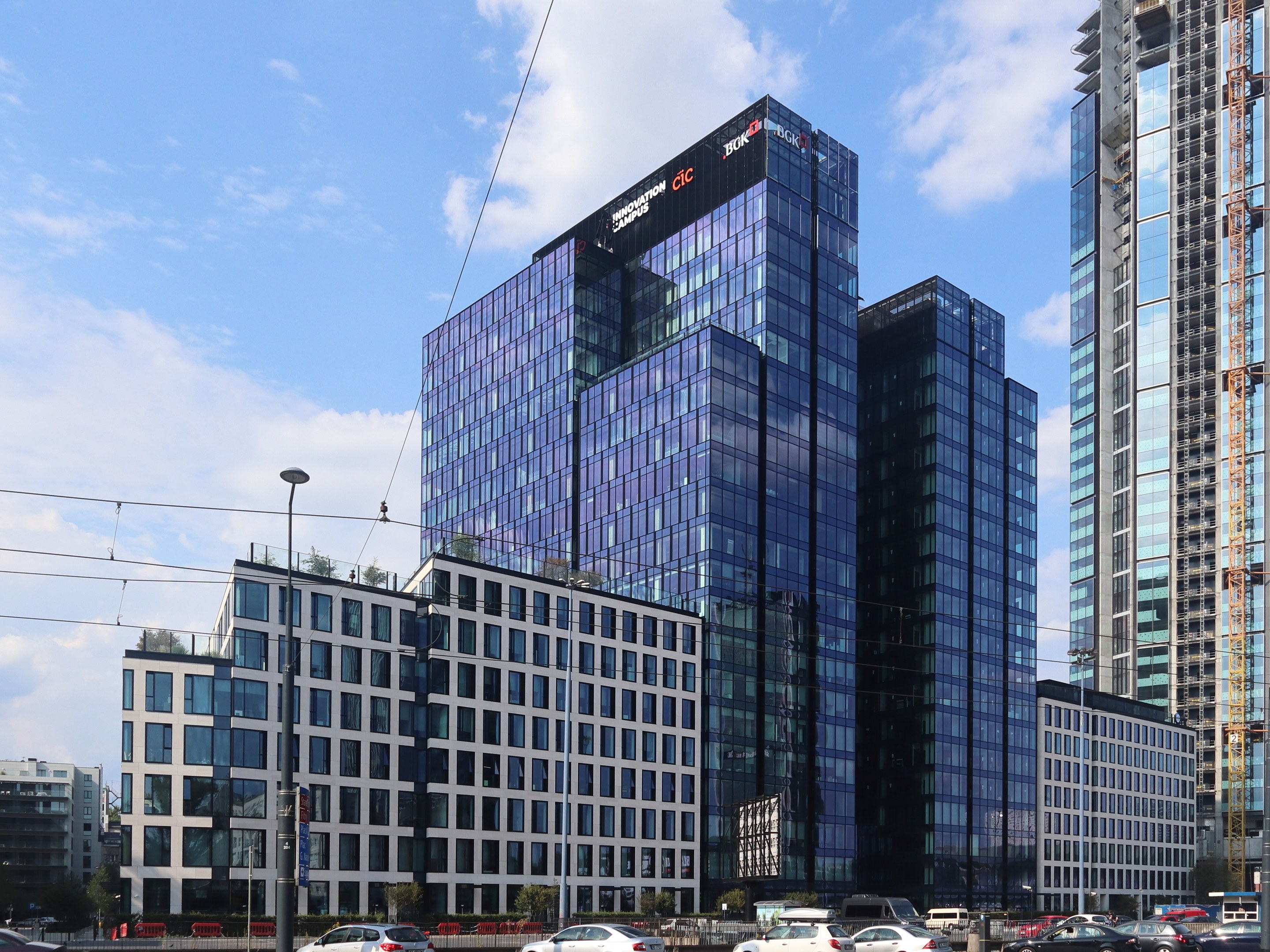 Außenansicht der Deutschen Bank Hauptstadquarters in Frankfurt, Deutschland, mit einem mehrstöckigen Gebäude mit zahlreichen Fenstern, umgeben von städtischer Infrastruktur wie Straßenlaternen, Fahrzeugen und Grünflächen unter einem bewölkten Himmel.