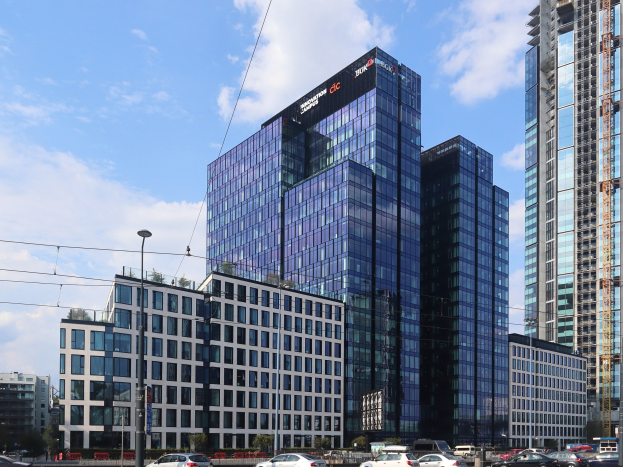 Außenansicht der Deutschen Bank Hauptstadquarters in Frankfurt, Deutschland, mit einem mehrstöckigen Gebäude mit zahlreichen Fenstern, umgeben von städtischer Infrastruktur wie Straßenlaternen, Fahrzeugen und Grünflächen unter einem bewölkten Himmel.