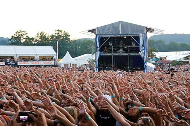Menschenmenge auf einem Musikfestival mit erhobenen Händen, einige halten Handys, vor einer Bühne mit Künstlern, weißen Zelten, Bäumen und einem klaren blauen Himmel.