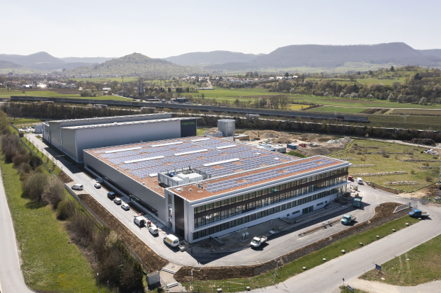 Luftaufnahme eines große Industriegebäudes mit Solarpanelen auf dem Dach, umgeben von Fahrzeugen, Bäumen, Gras und Strommasten; das erste solarbetriebene Werk in Deutschland.