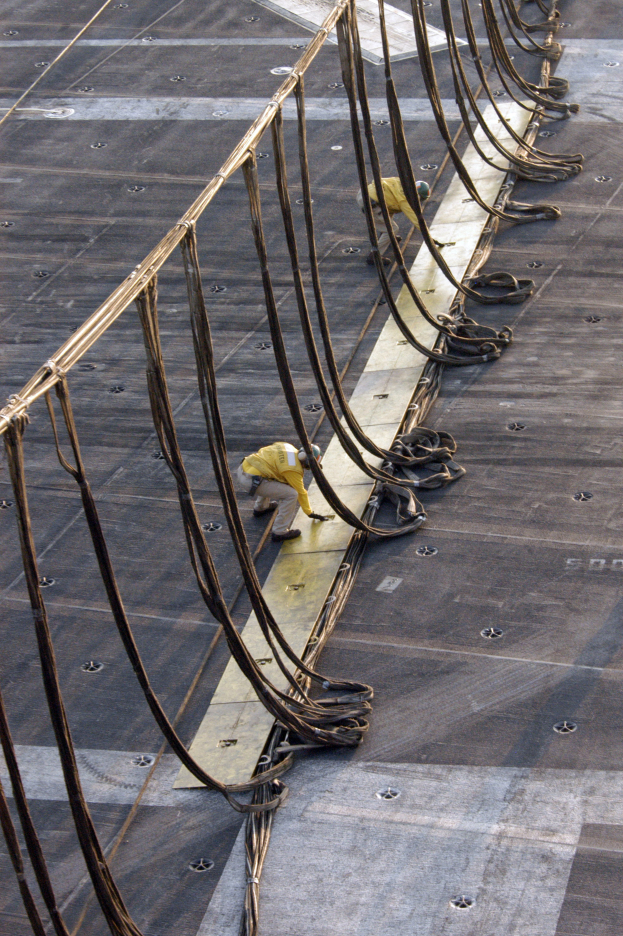 Zwei Männer in gelber Kleidung arbeiten an einer großen Metallbrückekonstruktion umgeben von Eisenstangen und Drähten.