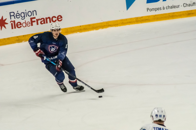 Ein Eishockeyspieler auf dem Eis mit einem Stock in der Hand, der eine Sportuniform und einen Helm trägt, mit einer textierten Wand im Hintergrund.
