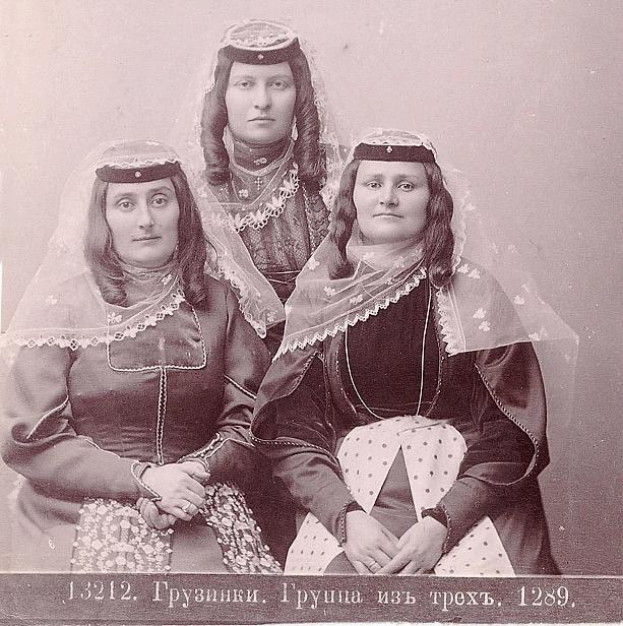 Drei Frauen in traditionellen russischen Hochzeitskleidern mit aufwendiger Stickerei und Perlen posieren vor einer weißen Wand in einem Schwarz-Weiß-Foto.