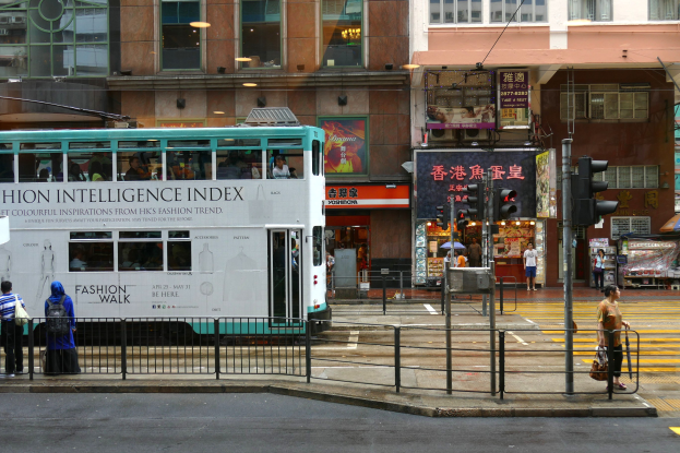 Ein Doppeldeckerbus fährt auf einer Straße mit hohen Gebäuden, Füßgängern auf dem Gehweg, einem Geländer, Verkehrsampeln und Informationsanzeigen.