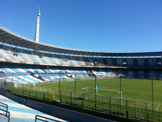Großes Stadion mit einem von einem Zaun umgebenen Fußballfeld, einen Turm im Hintergrund und einem klaren blauen Himmel.