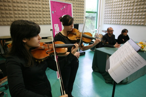 Zwei Frauen, die Geige spielen, ein Notenständer und ein Tisch im Vordergrund, sowie zwei Personen auf Stühlen rechts und eine Tafel im Hintergrund.