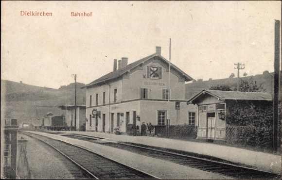 Schwarz-weiß-Fotografie eines Bahnhofs in Dielkirchen, Deutschland, mit einem Zug auf den Gleisen, umgeben von Häusern, Strommasten, Bäumen, Menschen und Hügeln im Hintergrund bei klarem Himmel.