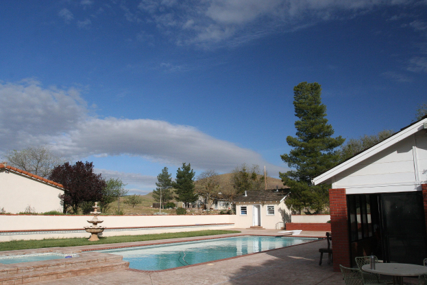 Schwimmbad mit einem zentralen Brunnen, umgeben von grünem Gras, Bäumen und Gebäuden unter einem klaren blauen Himmel, mit einem Tisch und Stühlen auf der rechten Seite.