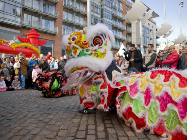 Ein lebendiges chinesisches Neujahrsfest in Amsterdam mit einer Löwendance-Vorführung vor einer Zuschauermenge, einige halten Kameras, vor einem Hintergrund aus Gebäuden, Laternenmästen und einem klaren blauen Himmel.