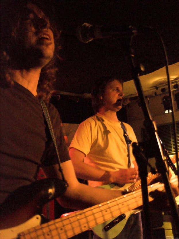 Eine Person in einem weißen Hemd singt in ein Mikrofon und spielt eine grüne Gitarre, während sie neben einer anderen Person in einem schwarzen Hemd, einer Brille und einer schwarzen Gitarre steht, mit Scheinwerfern und anderen Instrumenten im Hintergrund.