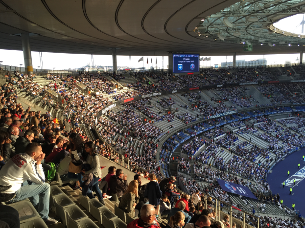 Eine große Menschenmenge sitzt im Allianz Stadion in München, Deutschland, und schaut ein Fußballspiel, mit einer Bühne auf der rechten Seite, Fahnen, Stangen und einem Bildschirm im Hintergrund und dem Himmel oben sichtbar.