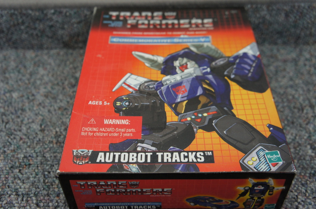 Ein roter, oranger und blauer Kasten mit einem Roboter und der Aufschrift "AUTOBOT TRACKS" oben drauf, vor einem unscharfen schwarzen Hintergrund.