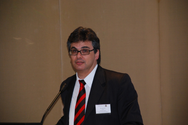 Eine Person in einem Anzug, die ein Abzeichen und eine Brille trägt, lächelt, während sie vor einem Mikrofon steht, das an einem Ständer befestigt ist, mit einer Wand im Hintergrund.