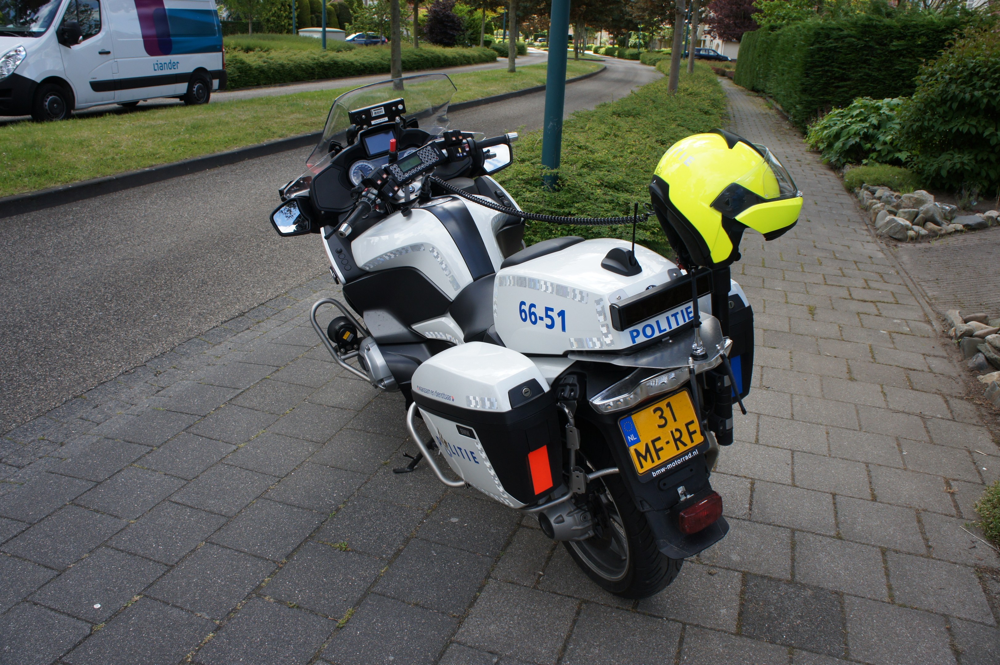 Ein Polizei-Motorrad am Straßenrand mit einem Helm darauf, umgeben von Bäumen, Polen, Gebäuden, Pflanzen, Gras, Steinen und anderen Fahrzeugen.