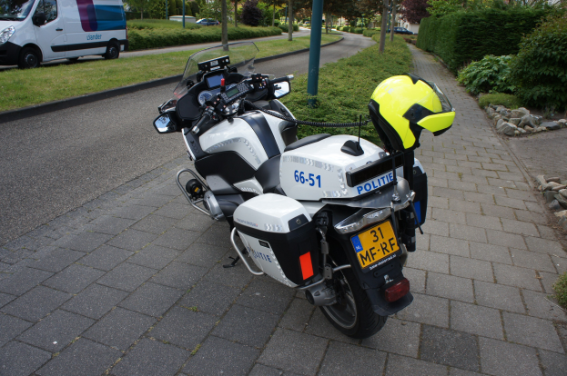 Ein Polizei-Motorrad am Straßenrand mit einem Helm darauf, umgeben von Bäumen, Polen, Gebäuden, Pflanzen, Gras, Steinen und anderen Fahrzeugen.