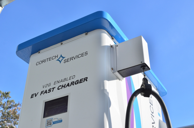 Elektrofahrzeug-Ladestation mit einem blauen und weißen "V2G Fast Charger"-Schild, Bäumen, einem Gebäude und einem klaren blauen Himmel.