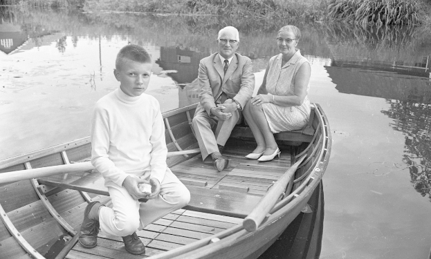 Schwarzes und wei├čes Foto von drei Menschen in einem Boot auf einem See, wobei einer ein wei├čes Kleid tr├Ągt und ein anderer ein Paddel h├Ąlt, umgeben von üppiger Vegetation und B├Ąumen.