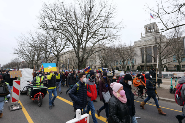 Eine große Gruppe von Menschen nimmt an einer Protestdemo auf einer Straße in Washington, D.C. teil, wobei einige Schilder und Plakate halten, andere Fahrräder fahren und Schilder, Bäume und ein klarer blauer Himmel im Hintergrund zu sehen sind.