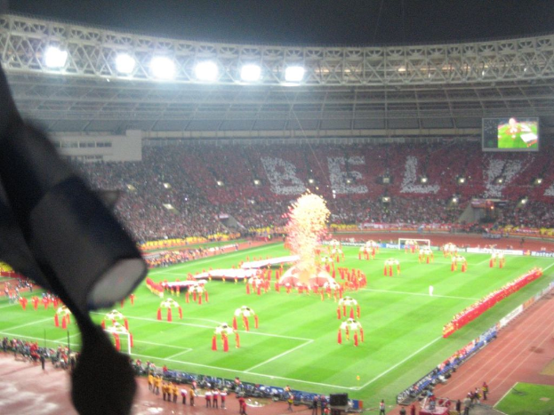 Ein Stadion mit Flutlicht, Eisengittern, Zuschauern, stehenden Personen, Werbetafeln, Anzeigetafeln, Sicherheitskameras, Flaggen und Deko.