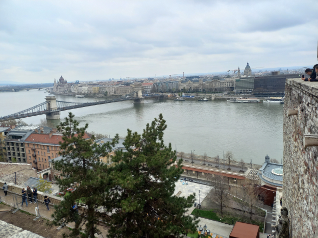 Ein Blick auf den Donaufluss von einem Hügel in Budapest, Ungarn, mit Menschen, Fahrzeugen, Gebäuden, einer Brücke, Booten, Kränen, Hügeln und einem bewölkten Himmel.