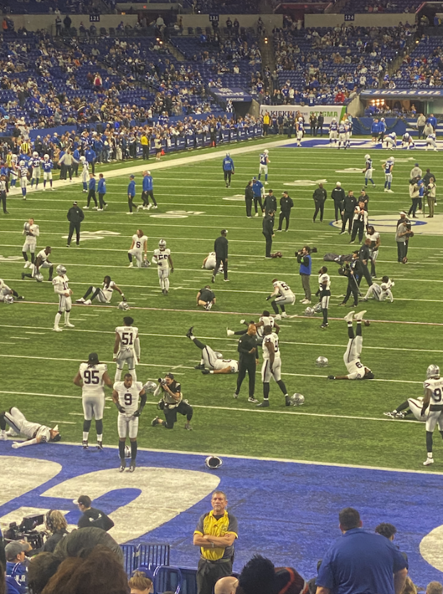 Ein Fußballspiel wird in einem Stadion mit Menschen auf dem Rasen und einigen auf Stühlen im Hintergrund gespielt, während eine Person eine Kamera hält, was auf eine Live-Übertragung hinweist.