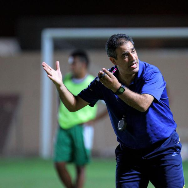 Ein Mann in einem blauen T-Shirt und Hose steht auf einem Fußballfeld und hält ein Mikrofon in der Hand, während er zu seinen Spielern gestikuliert. Im Hintergrund sind eine Person in einem grünen Kleid, ein Torpfosten und eine Wand zu sehen.