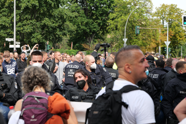 Eine große Gruppe von Menschen steht vor einer Menge von Polizeibeamten, einige tragen Masken und Rðcken, andere halten Kameras, mit Laternenmästen, Verkehrssignalen, Schildern, Bäumen und einem klaren blauen Himmel im Hintergrund.