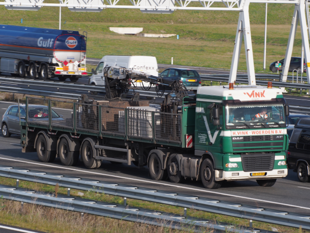 Ein großer Lastwagen mit Anhänger fährt auf einer Autobahn neben einer Brücke, mit Gras und Geländern auf beiden Seiten, unter einem klaren blauen Himmel mit Masten im Hintergrund.