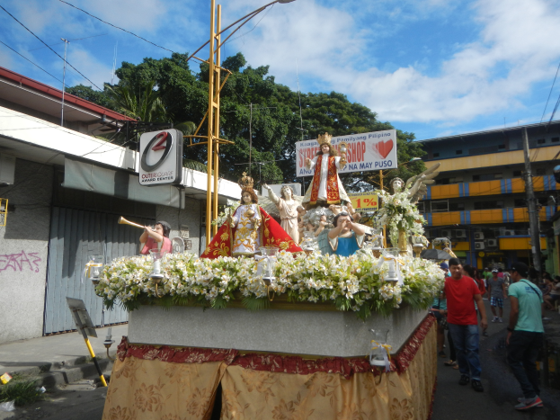 Ein Umzugswagen mit Statuen, Blumen und Texttafeln, der eine Straße mit Laternen, Gebäuden, Bäumen und einem bewölkten Himmel entlangfährt.
