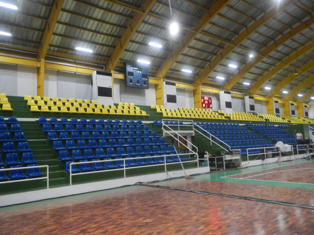 Großer Hallensportplatz mit Holzboden, blauen und gelben Sitzplätzen, Geländern, Treppen zum Sitzbereich, wandmontierten Bildschirmen, Deckenleuchten und einigen anwesenden Personen.