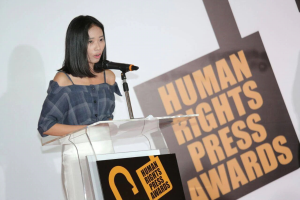 Eine Frau in einem schwarzen Kleid steht an einem Podium mit einem Mikrofon und hält eine Rede vor einem Banner, auf dem "Human Rights Press Awards" steht. Ihr Haar ist zurückgebunden und ihr Gesichtsausdruck ernst und konzentriert.