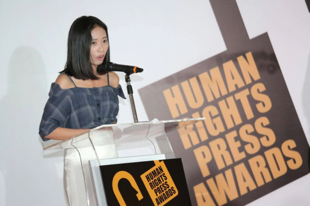 Eine Frau in einem schwarzen Kleid steht an einem Podium mit einem Mikrofon und hält eine Rede vor einem Banner, auf dem "Human Rights Press Awards" steht. Ihr Haar ist zurückgebunden und ihr Gesichtsausdruck ernst und konzentriert.