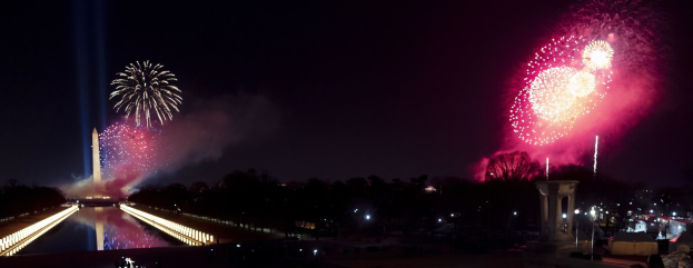 Nachthimmel erleuchtet von Feuerwerk über einem Gewässer, mit Bäumen, Pfählen, Lichtern und einem Turm im Hintergrund und Menschen, die sich um das Wasser versammeln.