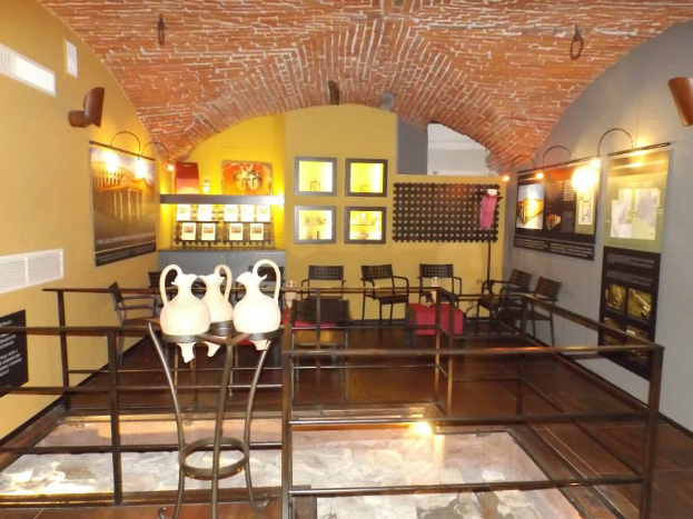 Ein Museumsraum voller Tische, Stühle, eines Geländers, Vasen, Texttafeln, Wandrahmen, Lampen und diverser Ausstellungsstücke.