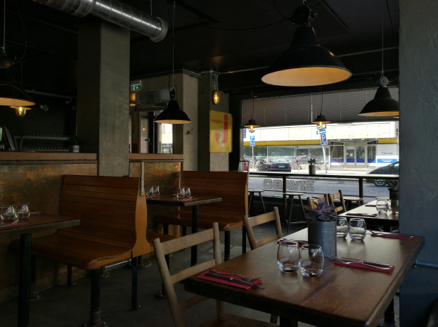 Ein Restaurant-Innenraum mit hölzernen Tischen und Stühlen, die mit Gläsern, Besteck, Servietten und einer Blumenvase gedeckt sind, beleuchtet von Hängelampen.