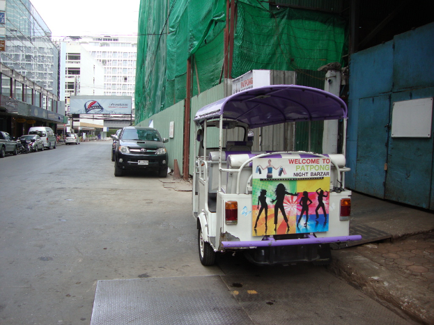 Gebäude, einige geparkte Autos, eine Werbetafel, ein bewölkter Himmel und ein Auto-Rikscha am Straßenrand.