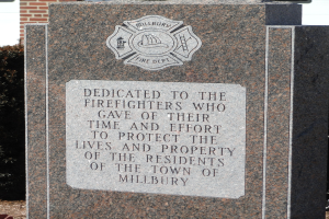 Denkmalstein mit einer Gedenktafel für Feuerwehrleute in Millbury, aufgestellt vor einem Backsteingebäude mit umliegender Vegetation.