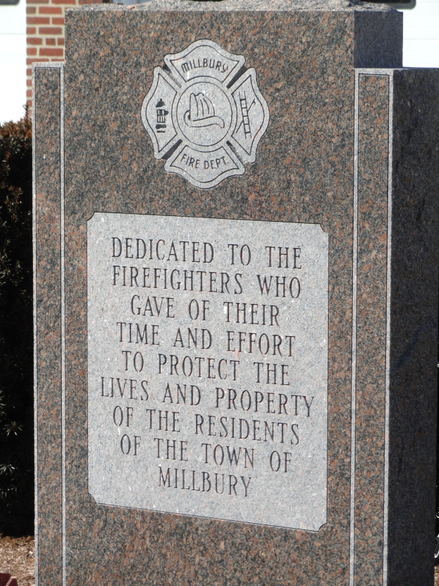 Denkmalstein mit einer Gedenktafel für Feuerwehrleute in Millbury, aufgestellt vor einem Backsteingebäude mit umliegender Vegetation.