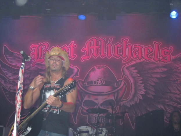 Eine Musikkonzert-Szene mit einem Gitarristen auf der Bühne, einer Person im unteren rechten Eck, einem Banner im Hintergrund und Drogen in der Mitte unten.