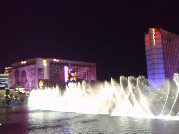 Ein Brunnen in der Mitte mit Wasser am Boden, umgeben von Gebäuden, Plakaten, Bildschirmen, Lichtern, Pflanzen und Himmel im Hintergrund.