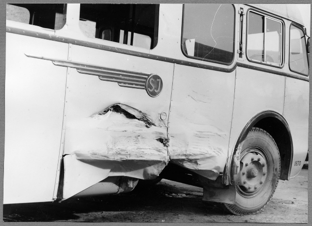 Ein Schwarz-Weiß-Foto eines beschädigten Busses am Straßenrand mit einer eingedrückten Front, zersplitterten Fenstern und einer offenen Fahrertür, die das Innere zeigt.