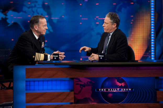 Zwei Männer in schwarzen Blazern, Hemden und Krawatten sitzen an einem Tisch mit Mikrofonen zwischen ihnen und sind in ein Gespräch vor einem Fernseher und einem blauen Hintergrund vertieft.