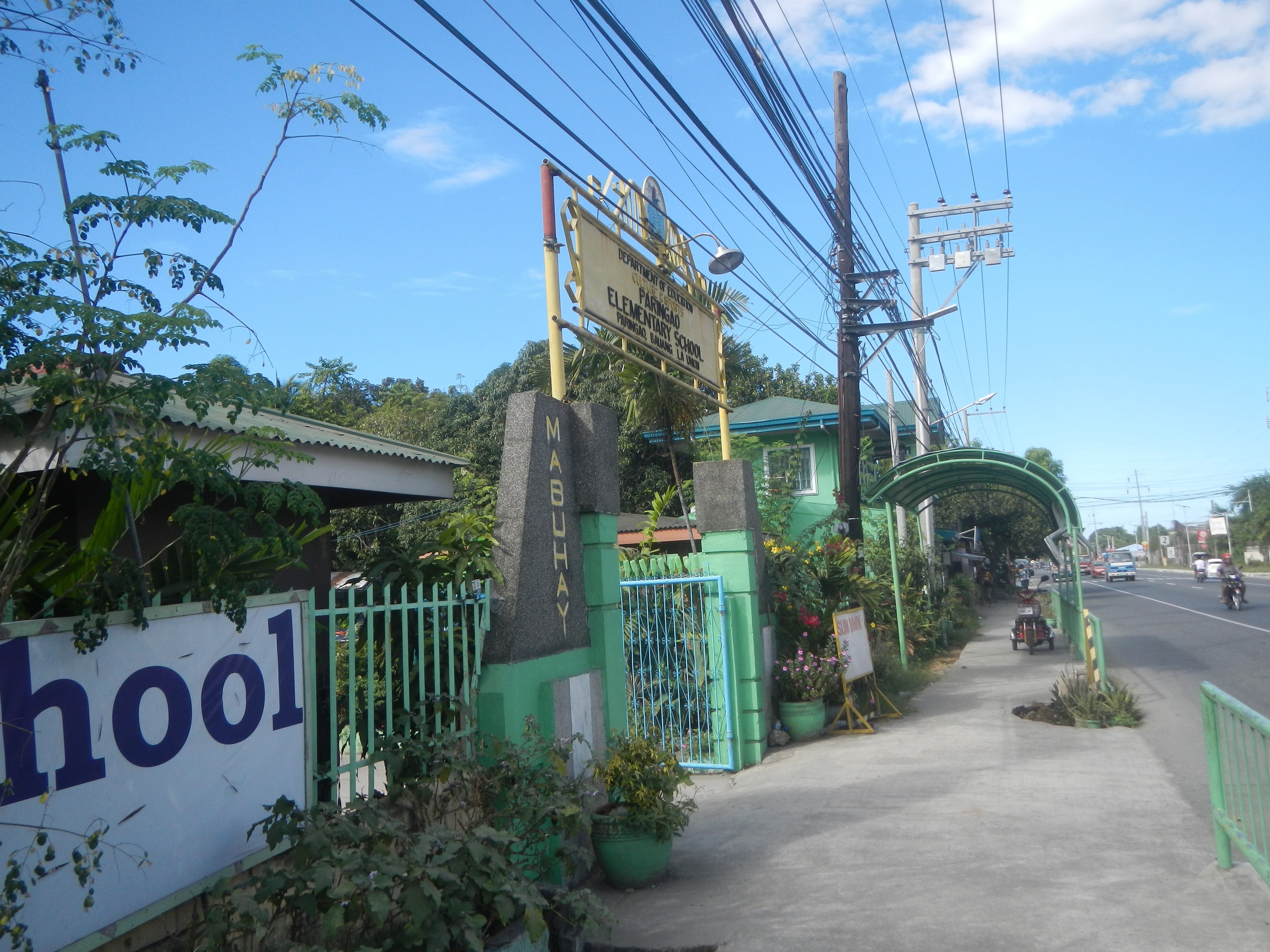 Außenansicht eines Schulgebäudes mit einem "Schule"-Schild, Fahrzeugen auf der Straße, einem Torbogen, einem Tor, einer Namenstafel, Grünfläche, Strommasten und einem bewölkten Himmel.
