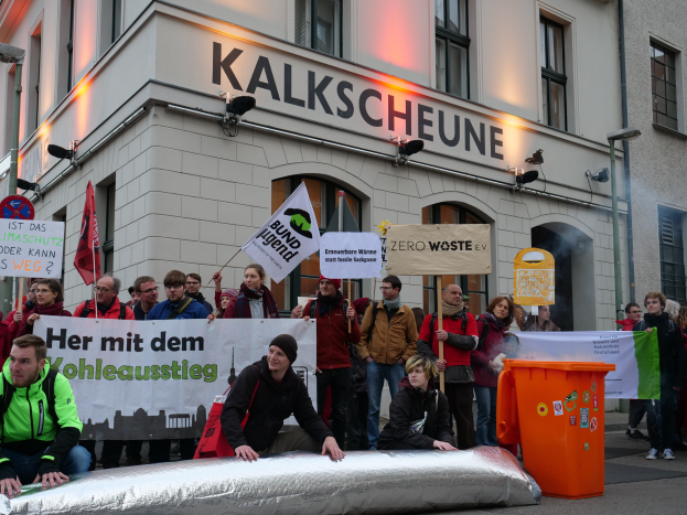 Eine Gruppe von Menschen mit Schildern und Plakaten steht vor einem Gebäude, mit zwei Personen im Vordergrund und einem Müllimer auf der rechten Seite, während einer Demonstration in Deutschland, mit Gebäuden, Fenstern, Lichtern und Schildern im Hintergrund.