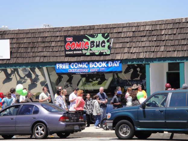 Gruppen von Menschen vor einem Gebäude mit Glastüren und einem Dach, zwei Autos auf der Straße, grüne Ballons, ein Banner und eine Namensplakette auf dem Dach, unter einem klaren Himmel.